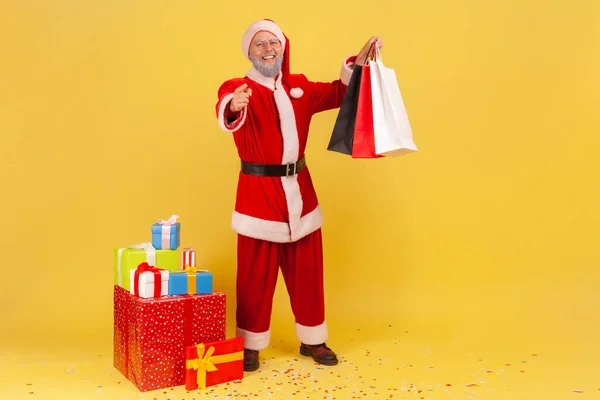 カメラを指して ショッピングバッグを保持サンタクラスの衣装を着て灰色のひげを持つ積極的な高齢者の男性は クリスマス休暇のプレゼント 黄色の背景に隔離された屋内スタジオショット — ストック写真