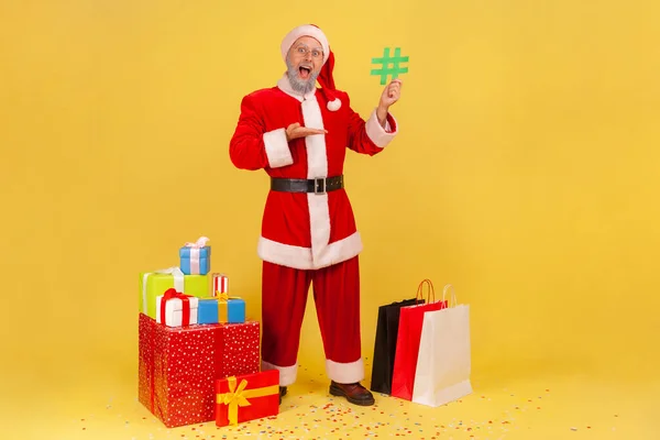 クリスマスプレゼントに囲まれたサンタのクラスの衣装を着てひげを生やした老人の完全な長さの肖像画は 彼の手の中に緑のハッシュタグを指しています 黄色の背景に隔離された屋内スタジオショット — ストック写真