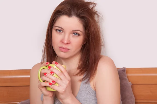 Sick woman in frustration. Closeup image of young woman with red nose holding a cup of tea while sitting in bed with copy space — Stock Photo, Image