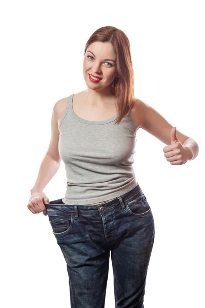 Retrato de larga duración de una atractiva joven y delgada mujer sonriente en pantalones vaqueros grandes que muestra una pérdida de peso exitosa con el pulgar hacia arriba, aislada sobre un fondo blanco —  Fotos de Stock