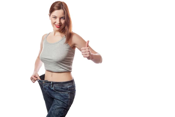 Full-length portrait of attractive slim young smiling woman in big jeans showing successful weight loss with her thumb up, isolated on white background — Stock Photo, Image