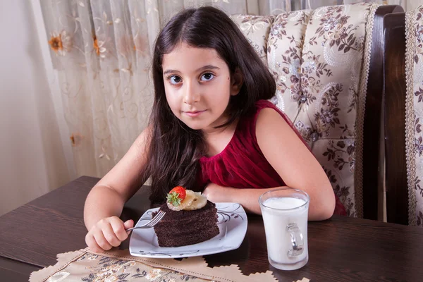Young small beautiful middle eastern child girl with chocolate cake with pineapple, strawberry, and milk with red dress and dark eyes and long hair happy drinking and eating at home and sitting smiling. looking at camera. studio shot. — Stock fotografie