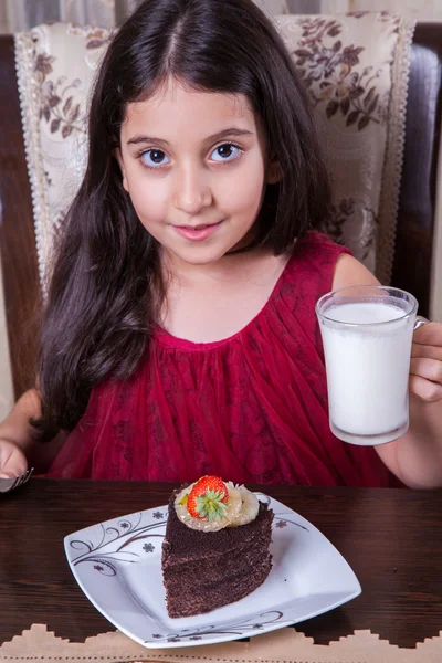 Joven niña pequeña y hermosa de Oriente Medio con pastel de chocolate con piña, fresa y leche con vestido rojo y ojos oscuros y cabello largo feliz bebiendo y comiendo en casa y sentado sonriendo. mirando a la cámara. plano de estudio . —  Fotos de Stock