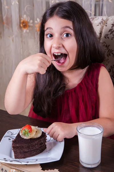 Joven niña pequeña y hermosa de Oriente Medio con pastel de chocolate con piña, fresa y leche con vestido rojo y ojos oscuros y cabello largo feliz bebiendo y comiendo en casa y sentado sonriendo. mirando a la cámara. plano de estudio . —  Fotos de Stock