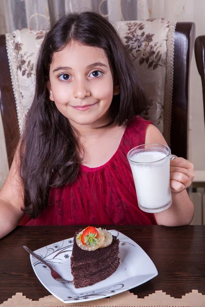 Joven niña pequeña y hermosa de Oriente Medio con pastel de chocolate con piña, fresa y leche con vestido rojo y ojos oscuros y cabello largo feliz bebiendo y comiendo en casa y sentado sonriendo. mirando a la cámara. plano de estudio . —  Fotos de Stock