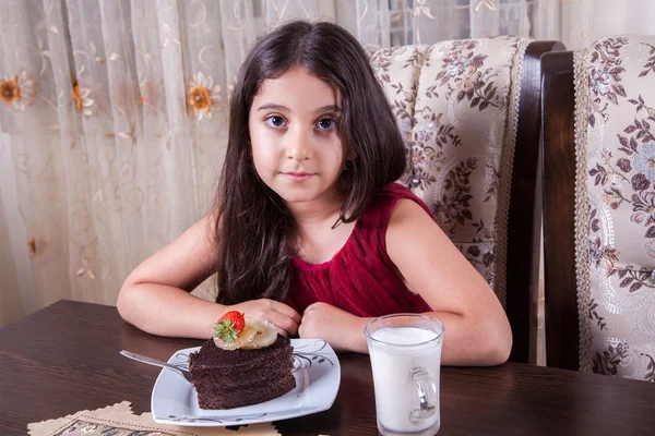 Joven niña pequeña y hermosa de Oriente Medio con pastel de chocolate con piña, fresa y leche con vestido rojo y ojos oscuros y cabello largo feliz bebiendo y comiendo en casa y sentado sonriendo. mirando a la cámara. plano de estudio . —  Fotos de Stock