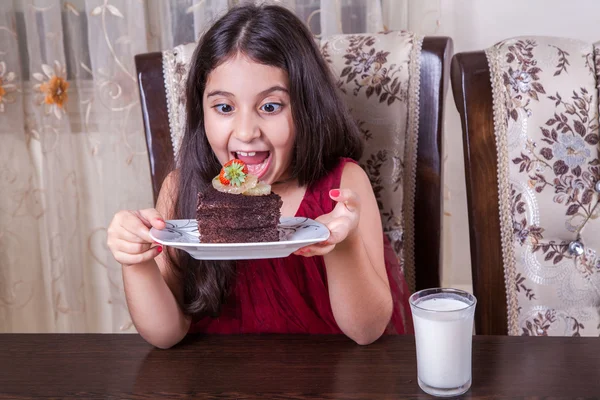 Joven niña pequeña y hermosa de Oriente Medio con pastel de chocolate con piña, fresa y leche con vestido rojo y ojos oscuros y cabello largo feliz bebiendo y comiendo en casa y sentado sonriendo. mirando a la cámara. plano de estudio . —  Fotos de Stock