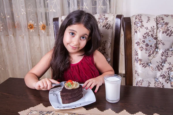 Joven niña pequeña y hermosa de Oriente Medio con pastel de chocolate con piña, fresa y leche con vestido rojo y ojos oscuros y cabello largo feliz bebiendo y comiendo en casa y sentado sonriendo. mirando a la cámara. plano de estudio . —  Fotos de Stock