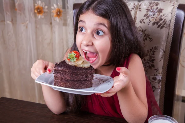 Young small beautiful middle eastern child girl with chocolate cake with pineapple, strawberry, and milk with red dress and dark eyes and long hair happy drinking and eating at home and sitting smiling. looking at camera. studio shot. — Stock Photo, Image