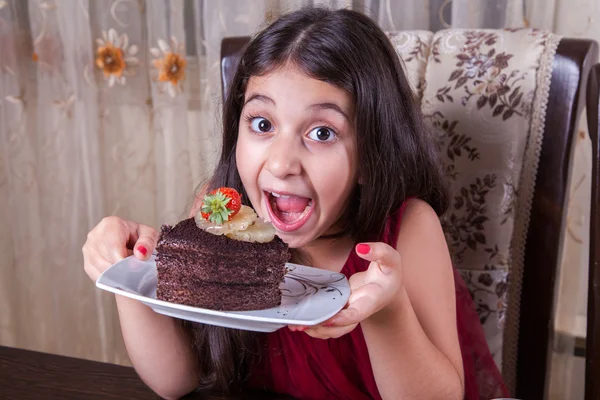 Joven niña pequeña y hermosa de Oriente Medio con pastel de chocolate con piña, fresa y leche con vestido rojo y ojos oscuros y cabello largo feliz bebiendo y comiendo en casa y sentado sonriendo. mirando a la cámara. plano de estudio . — Foto de Stock