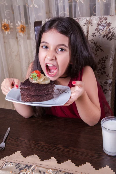 Young small beautiful middle eastern child girl with chocolate cake with pineapple, strawberry, and milk with red dress and dark eyes and long hair happy drinking and eating at home and sitting smiling. looking at camera. studio shot. — Stock Photo, Image