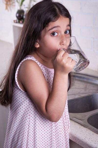 One beautiful middle eastern little girl with pink dress and long dark brown hair and eyes on white kitchen,helping parents to wash dishes and drinking water and smiling looking at camera. — Stock Photo, Image