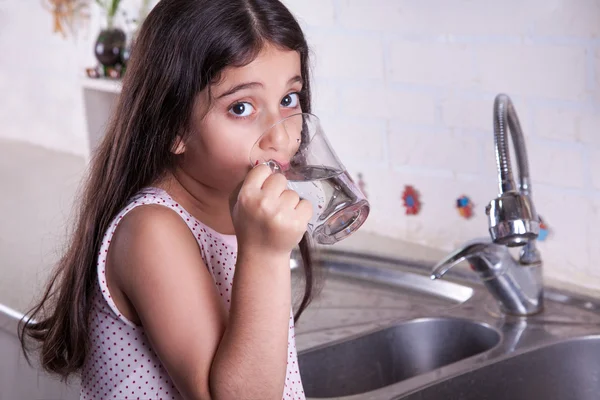 One beautiful middle eastern little girl with pink dress and long dark brown hair and eyes on white kitchen,helping parents to wash dishes and drinking water and smiling looking at camera. — Stock Photo, Image