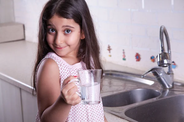 One beautiful middle eastern little girl with pink dress and long dark brown hair and eyes on white kitchen,helping parents to wash dishes and drinking water and smiling looking at camera. — Stock Photo, Image