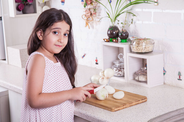 Beautiful little middle eastern 7 years old girl is working with knife and onion in the white kitchen.