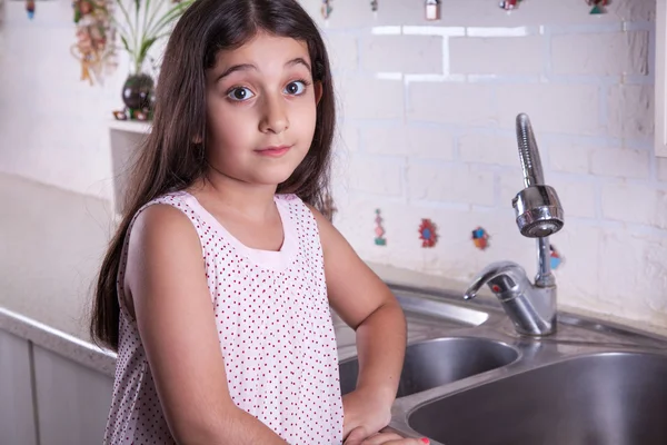 One beautiful middle eastern little girl with pink dress and long dark brown hair and eyes on white kitchen,helping parents to wash dishes and drinking water and smiling looking at camera. — Stock fotografie