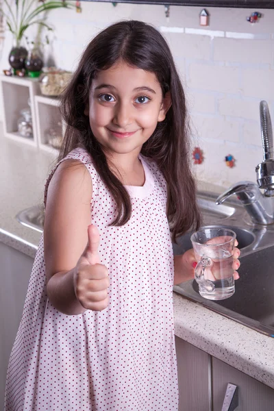 Una hermosa niña del medio oriente con vestido rosa y cabello largo de color marrón oscuro y ojos en la cocina blanca, ayudando a los padres a lavar los platos y beber agua y sonriendo mirando a la cámara . —  Fotos de Stock