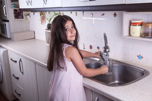 One beautiful middle eastern little girl with pink dress and long dark brown hair and eyes on white kitchen,helping parents to wash dishes and drinking water and smiling looking at camera. — Stock Photo, Image