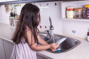 One beautiful middle eastern little girl with pink dress and long dark brown hair and eyes on white kitchen,helping parents to wash dishes and drinking water and smiling looking at camera.