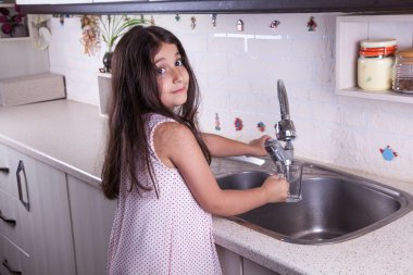 One beautiful middle eastern little girl with pink dress and long dark brown hair and eyes on white kitchen,helping parents to wash dishes and drinking water and smiling looking at camera.