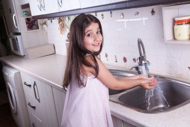 One beautiful middle eastern little girl with pink dress and long dark brown hair and eyes on white kitchen,helping parents to wash dishes and drinking water and smiling looking at camera.