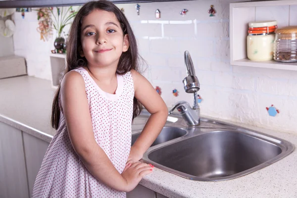 One beautiful middle eastern little girl with pink dress and long dark brown hair and eyes on white kitchen,helping parents to wash dishes and drinking water and smiling looking at camera. — Stock fotografie