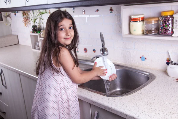 One beautiful middle eastern little girl with pink dress and long dark brown hair and eyes on white kitchen,helping parents to wash dishes and drinking water and smiling looking at camera.