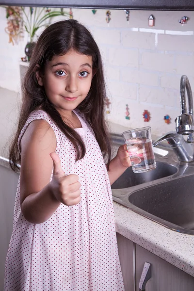 One beautiful middle eastern little girl with pink dress and long dark brown hair and eyes on white kitchen,helping parents to wash dishes and drinking water and smiling looking at camera. — Stock Photo, Image