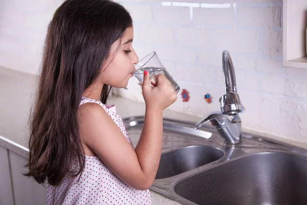 One beautiful middle eastern little girl with pink dress and long dark brown hair and eyes on white kitchen,helping parents to wash dishes and drinking water and smiling looking at camera. — Stockfoto