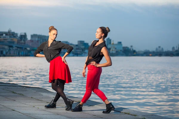 Twee jonge mooie tweelingzusjes dansen waacking dans in de achtergrond van de stad in de buurt van de rivier. — Stockfoto