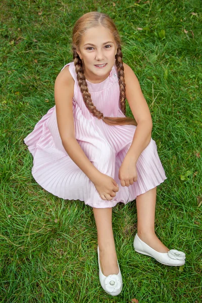 Retrato de uma menina sorridente sentada na grama verde com sorriso de dente e estilo de cabelo pigtail olhando para a câmera e feliz. vista superior. 8-11 anos . — Fotografia de Stock