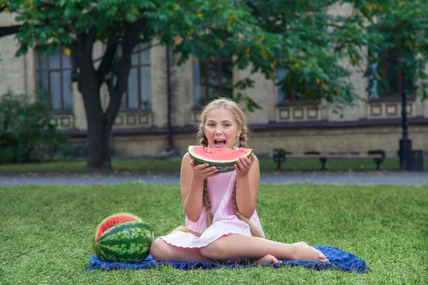 Linda niña comiendo sandía en la hierba en verano. con el pelo largo de cola de caballo y sonrisa dentada sentado en la hierba y disfrutando . — Foto de Stock