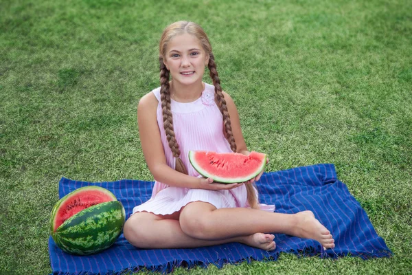 Linda niña comiendo sandía en la hierba en verano. con el pelo largo de cola de caballo y sonrisa dentada sentado en la hierba y disfrutando . — Foto de Stock