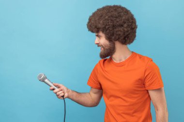 Side view of man with Afro hairstyle wearing orange T-shirt standing offering microphone, journalist taking interview, looking ahead. Indoor studio shot isolated on blue background.