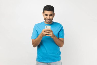 Portrait of positive smiling unshaven man blogger wearing blue T- shirt standing using smart phone, checking social networks, writing post. Indoor studio shot isolated on gray background.