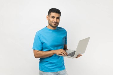 Portrait of smiling satisfied attractive unshaven man wearing blue T- shirt standing holding laptop in hands, working online, looking at camera. Indoor studio shot isolated on gray background.