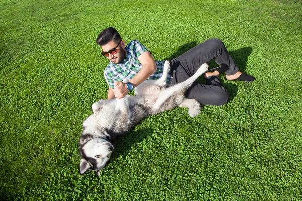 Un hombre de la moda de Oriente Medio con barba y estilo de pelo de moda está descansando y disfrutar de la hermosa hierba verde día y jugar con su perro Hasky. mirando a la cámara con sonrisa y felicidad . — Foto de Stock
