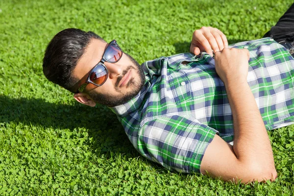 One fashion middle eastern man with beard, fashion hair style is resting on beautiful green grass day time. young arab businessman 20-30 years, resting after hard work. smart phone, tablet.