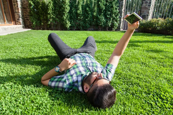 Un joven hombre de la moda de Oriente Medio con barba y estilo de pelo de moda está acostado en una hierba en un parque tomando selfie. sosteniendo el teléfono inteligente, tableta, cámara y mirando a la cámara . —  Fotos de Stock