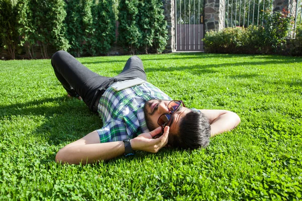 Un uomo medio orientale di moda con la barba, stile di capelli di moda sta riposando su bella erba verde giorno. giovane uomo d'affari arabo 20-30 anni, a riposo dopo un duro lavoro. smartphone, tablet . — Foto Stock