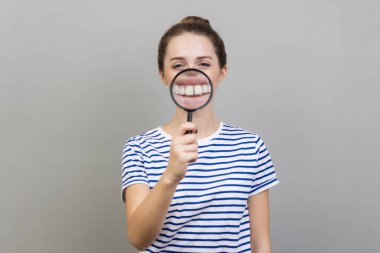 Çizgili tişört giyen komik bir kadının portresi. Dişlerinin üzerinde büyüteç olan, dişlek bir gülümsemeyle bakan. Gri arka planda izole edilmiş kapalı stüdyo görüntüleri.