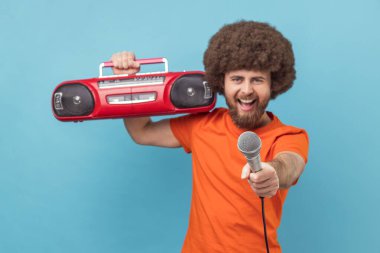 Turuncu tişörtlü, Afro saçlı, omzunda kırmızı bant kayıt cihazı olan ve müzik hakkında fikir soran mutlu bir adam. Mavi arka planda kapalı stüdyo çekimi.