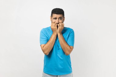 Mavi tişört giyen, hayal kırıklığına uğramış, tıraşsız bir adamın portresi ayakta duran tırnak ısıran, gergin ifadeli, korku dolu gözlerle gözlerini kaçıran. Gri arka planda izole edilmiş kapalı stüdyo görüntüleri.