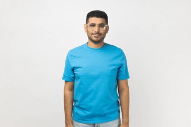 Mavi tişört giyen, optik gözlüklü, pozitif ifade ile kameraya bakan pozitif, tıraşsız bir adamın portresi. Gri arka planda izole edilmiş kapalı stüdyo görüntüleri.