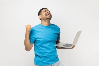 Mavi tişört giyen, dizüstü bilgisayarı elinde tutan, yumruğunu sıkan, bahis oynayan ve kazanan, başarıyı kutlayan son derece mutlu bir adamın portresi. Gri arka planda izole edilmiş kapalı stüdyo görüntüleri