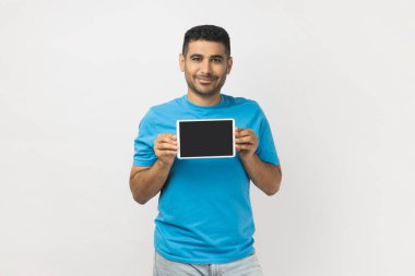 Mavi tişört giyen, siyah banka ekranı ve reklam alanı olan tablet gösteren iyimser, tatmin olmamış bir adamın portresi. Gri arka planda izole edilmiş kapalı stüdyo görüntüleri.