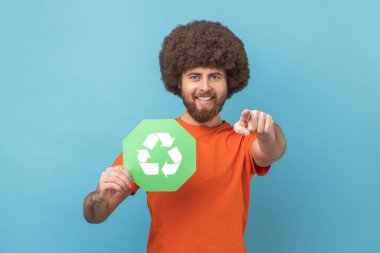 Turuncu tişörtlü Afro saçlı bir adamın portresi elinde atık geri dönüşüm sembolü, ekoloji hakkında endişelenen, kamerayı işaret eden. Mavi arka planda kapalı stüdyo çekimi.