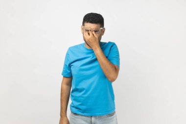 Mavi tişört giyen yorgun ve traşsız bir adamın portresi gözlüğünü çıkarıyor, gözlerini ovuyor, acıyı hissediyor, çok çalışıyor. Gri arka planda izole edilmiş kapalı stüdyo görüntüleri.