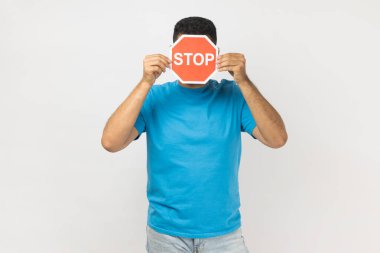 Portrait of unknown anonymous man wearing blue T- shirt standing hiding his face with behind red stop sign, prohibition, forbidden action. Indoor studio shot isolated on gray background.