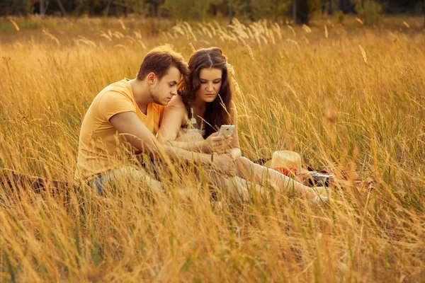 Liebespaar legt sich auf Blumenwiese im herbstlichen Park, warmer, sonniger Tag, genießt Familie, romantisches Date, Glück und Liebeskonzept. Halten und auf das Smartphone schauen. — Stockfoto